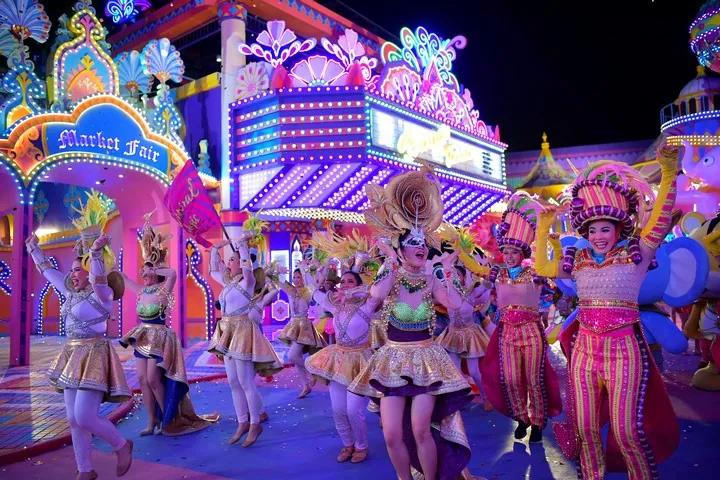 Des danseurs en costumes à paillettes et coiffes à plumes défilant devant des façades illuminées de néons à Carnival Magic, Phuket